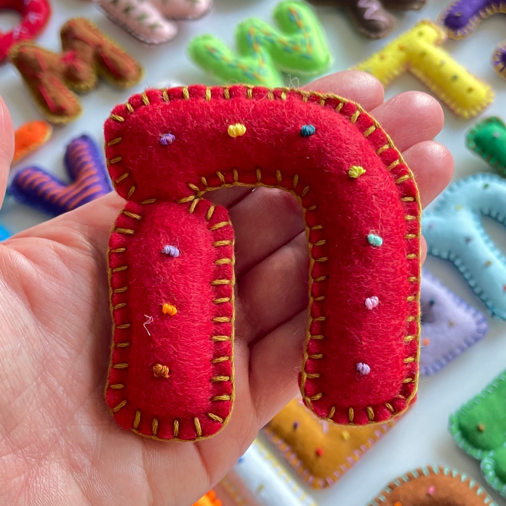Hand holding a red letter 'N' shaped fabric toy with colorful details against a background of various letter-shaped toys.