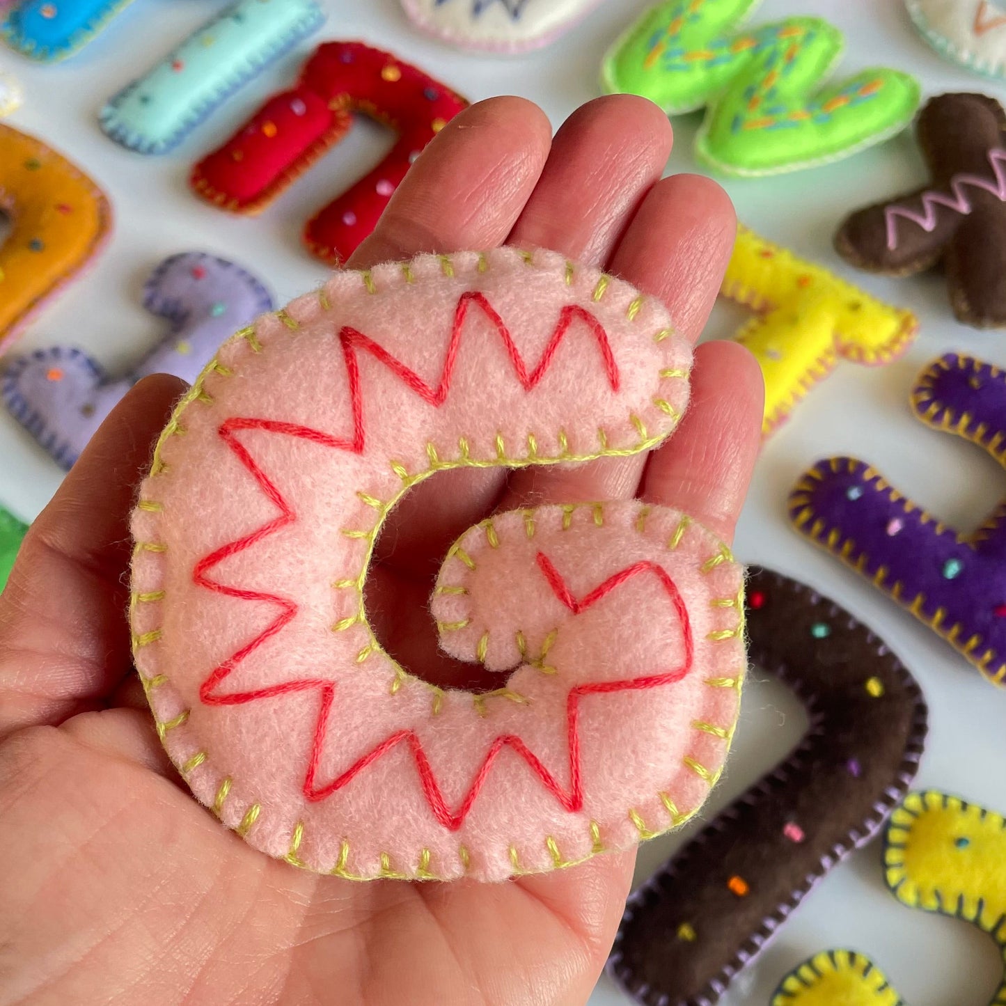 Hand holding a felt letter 'G' with red stitching against a colorful background of handmade felt letters.