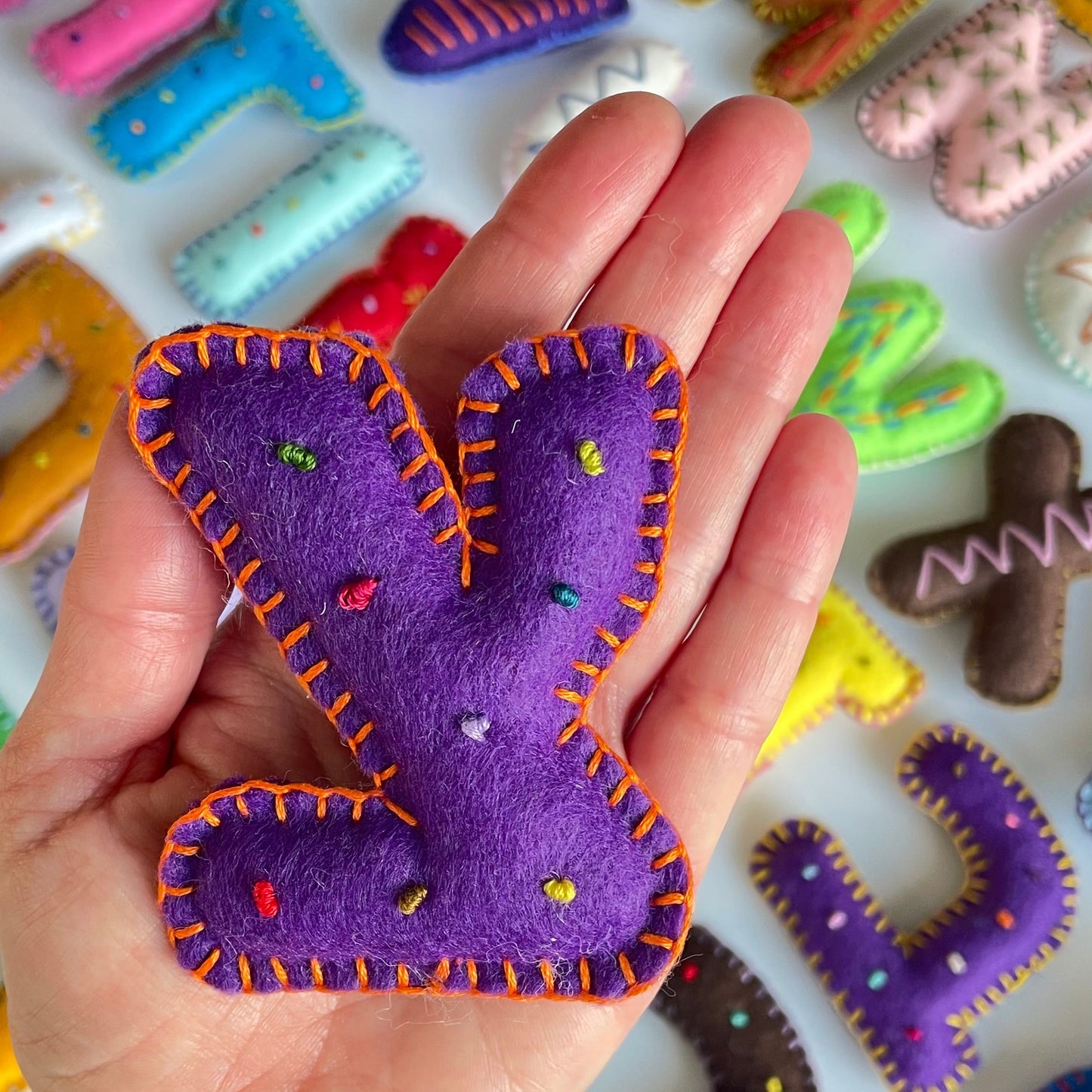 Hand holding purple felt Hebrew handmade embroidered letter Tzadi with colorful edges against a background of various felt letters.