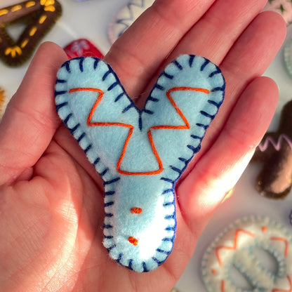 Hand holding a blue Y letter shaped felt ornament with orange accents against a colorful background of similar ornaments.