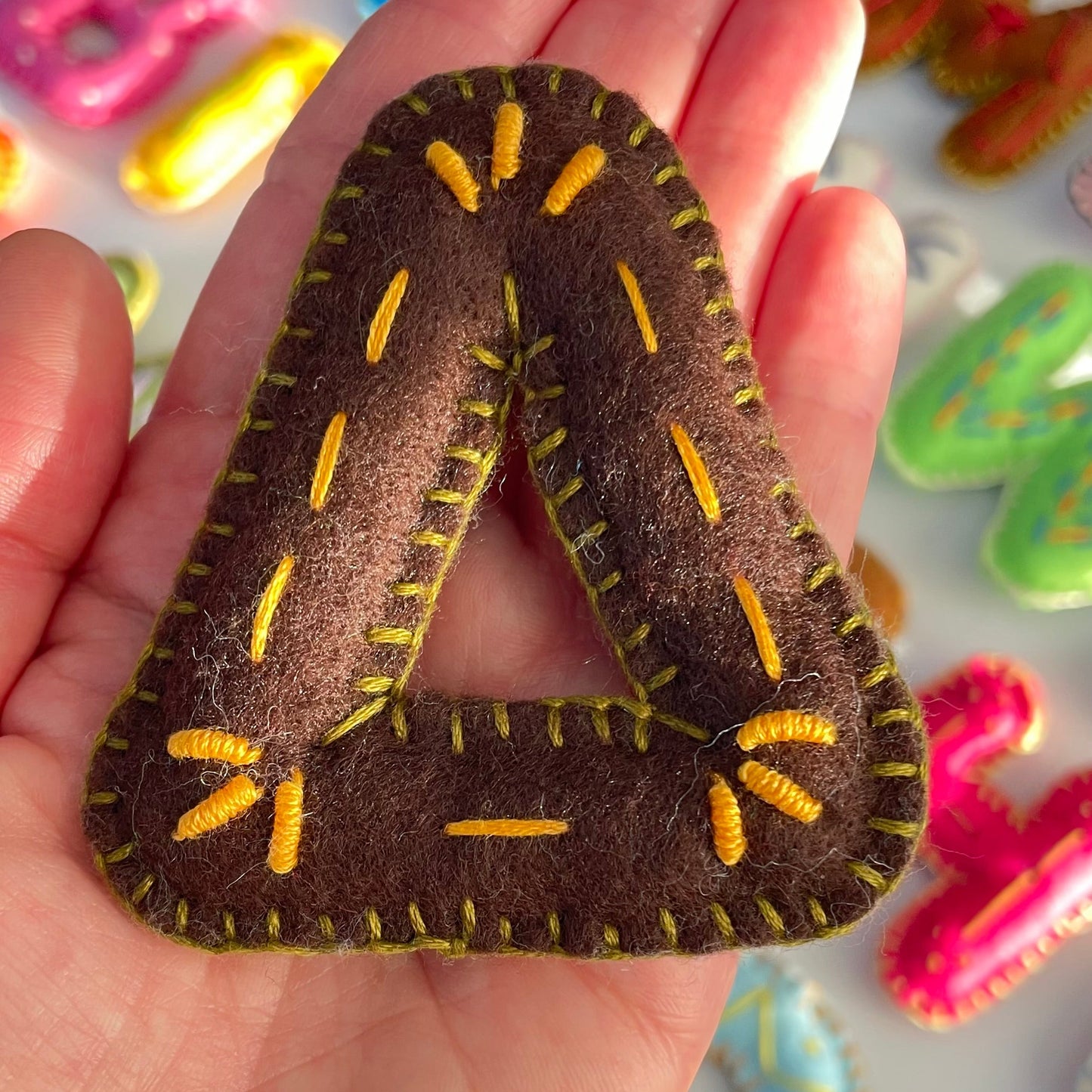Hand holding handmade greek Delta felt letter  with yellow stitching against a colorful background of felt letters.